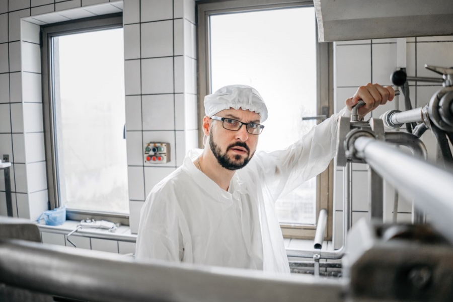 Ein Mann in weißem Laborkittel und Haarnetz  steht in einem gefliesten Raum. Er hält ein Verschlussventil in der Hand und blickt in die Kamera. Zwei Fenster sieht man im Hintergrund.